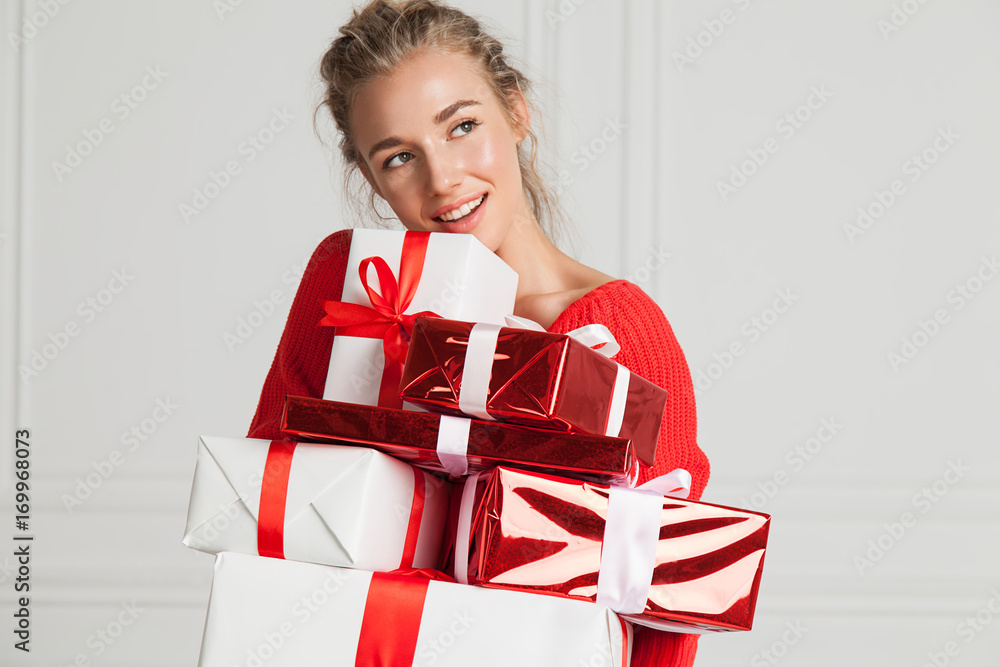 Fototapeta premium Close-up face of a beautiful smiling woman with a bunch of presents in a festive red sweater in a white interior