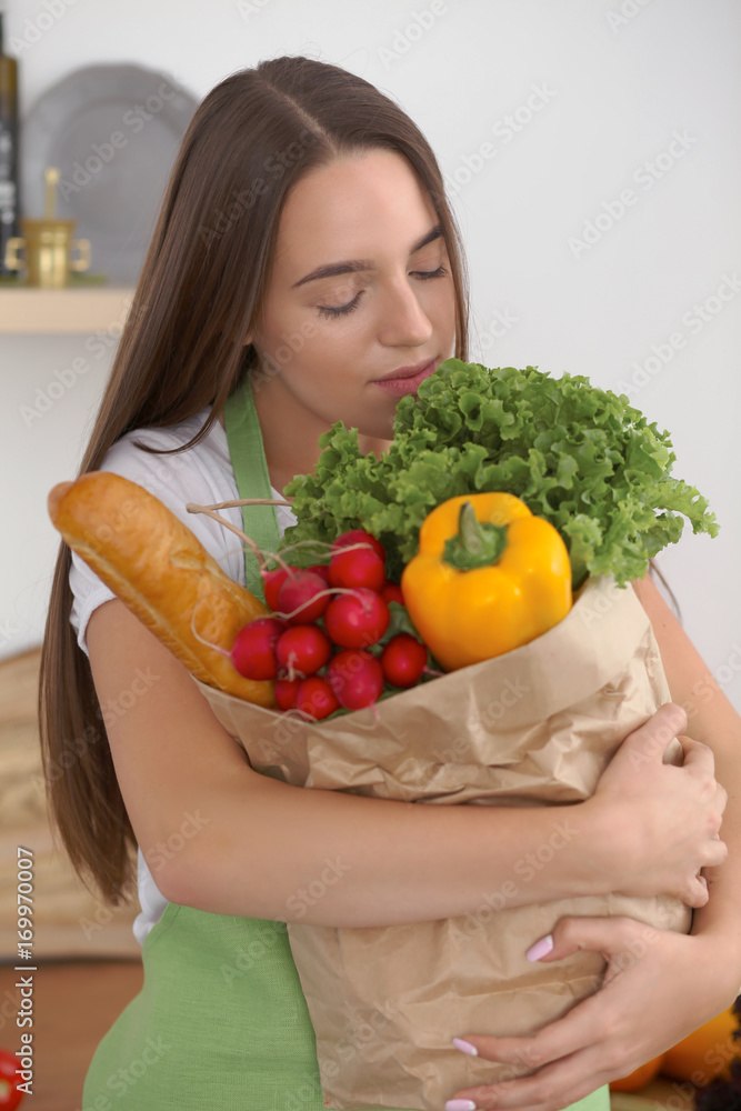 Young caucasian woman in a green apron is holding paper bag full of vegetables and fruits while smiling in kitchen Young caucasian woman in a green apron is holding paper bag full of vegetables and fruits while smiling in kitchen