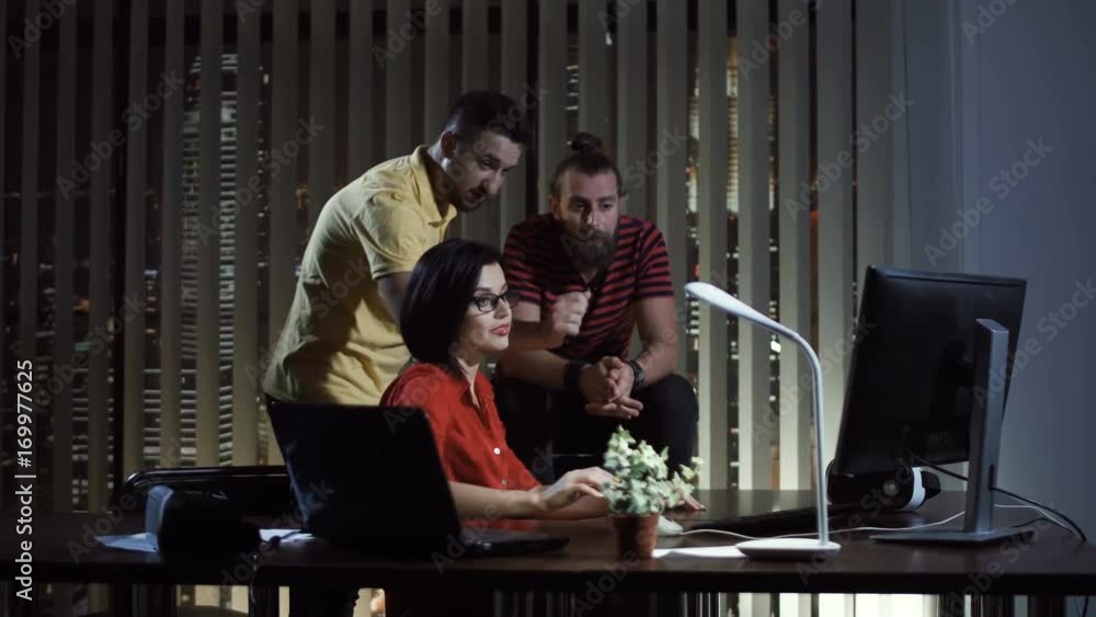 Smiling man and woman showing computer with work results to colleague in office late at night. Smiling man and woman showing computer with work results to colleague in office late at night.