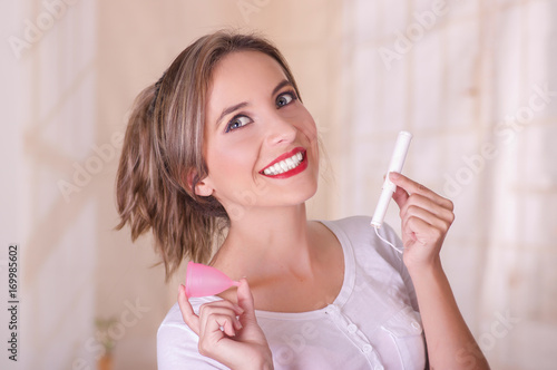 Close up of young beautiful smiling woman holding a menstruation cotton tampon in one hand and with her other hand a menstrual cup in a blurred background