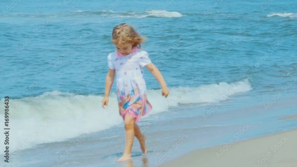 Adorable little girl  in a dress runs  jumping on the beach.  Child enjoys playing on the seashore running on the water with pleasure. splashing in sea waves. Stock ビデオ | Adobe Stock