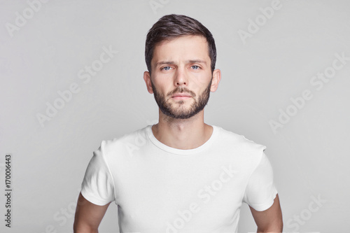 Close up portrait of good-looking serious bearded Caucasian man with blue beautiful eyes wearing white casual t-shirt posing isolated on gray studio wall with copy space for your promotional content.