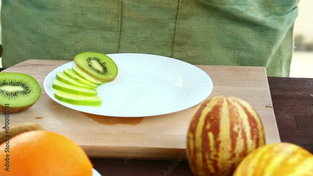 A woman puts pieces of kiwi, which is on a cutting board, on a plate. A woman puts pieces of kiwi, which is on a cutting board, on a plate.