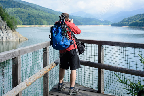 Photographer in a red jacket with a blue backpack on the lake. Lake Vidraru Carpathians Romania.