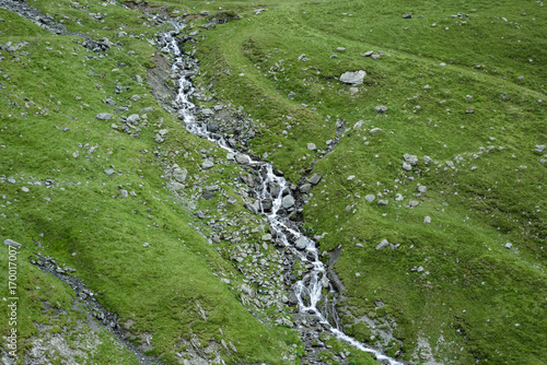 The flow takes place in the mountains through the way through the stones against the backdrop of green grass. Water, streams, waterfalls. 