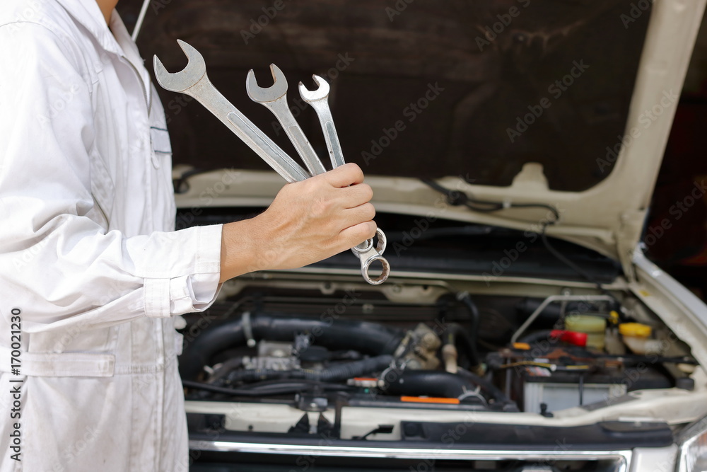 Portrait of professional young mechanic man in white uniform holding wrenches against car in open hood at the repair garage. Portrait of professional young mechanic man in white uniform holding wrenches against car in open hood at the repair garage.