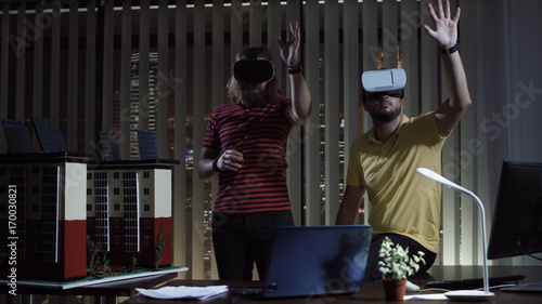 Two men using virtual reality helmet in office in the evening. Its two architects which design mockup of new eco building with solar panels on the roof.