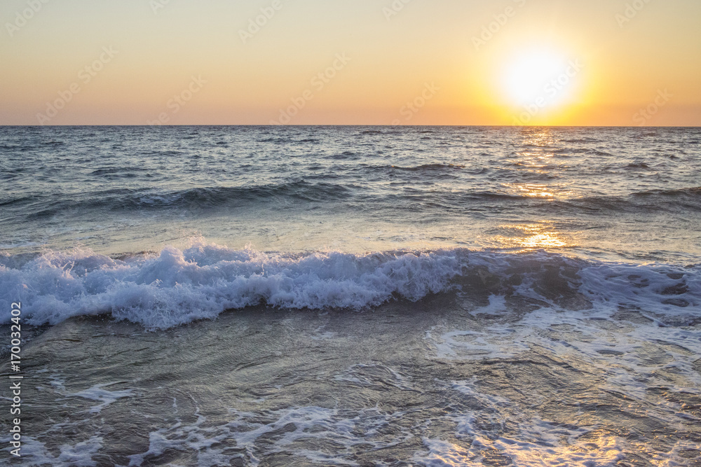 Panorama del sole che tramonta a mare. L'acqua è mossa e le onde a contatto con la sabbia formano la schiuma. Il mare è agitato e il cielo è arancione per via del crepuscolo.