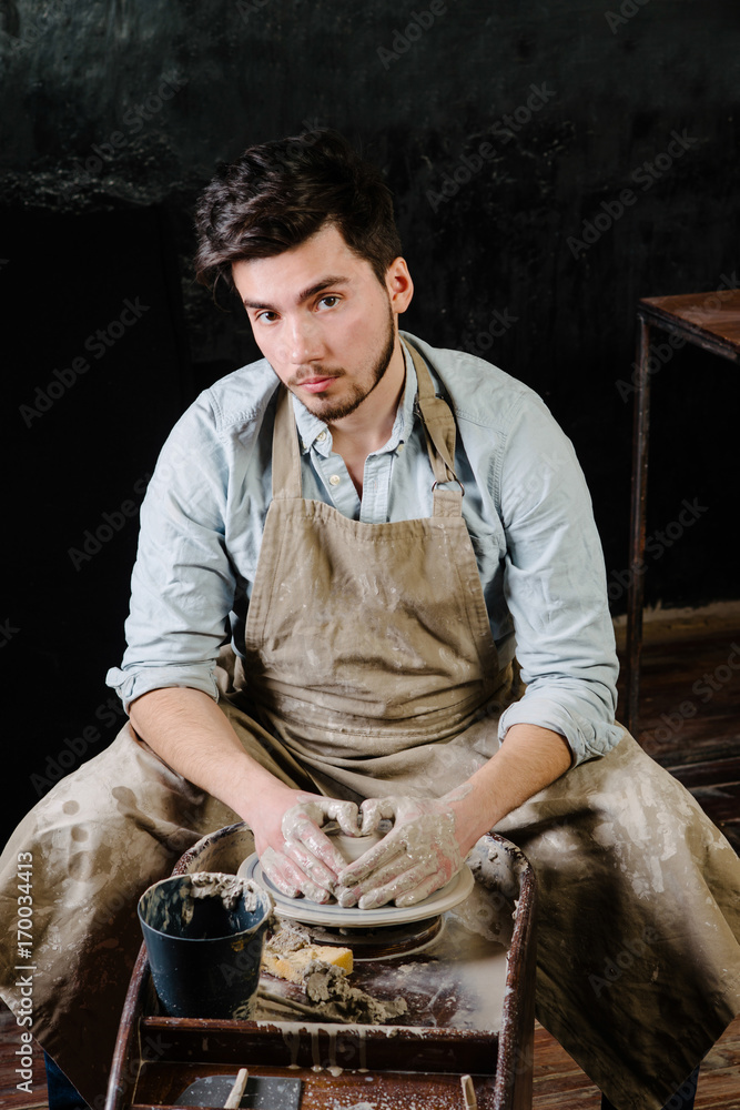 pottery, workshop, ceramics art concept - man working on potter's wheel with raw clay with hands, a male brunette sculpt a utensils near wooden table with tools, master in apron and a shirt pottery, workshop, ceramics art concept - man working on potter's wheel with raw clay with hands, a male brunette sculpt a utensils near wooden table with tools, master in apron and a shirt