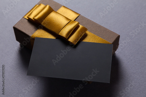 Blank black business card on gray table. Little gift box brown color with golden ribbon. Place for signature. Copy space. Corporate gift concept. Happy birthday concept.