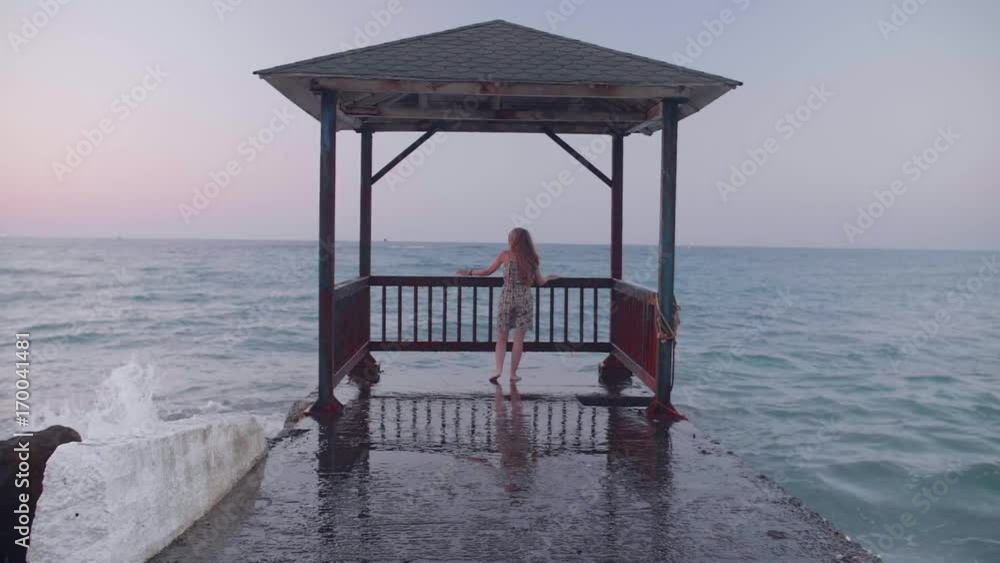 Young beautiful woman walking on pier in midi dress at sunset. Attractive blond girl standing near storm waves hitting the pier. Waves splashes in slow motion. Freedom,solitude concept. Young beautiful woman walking on pier in midi dress at sunset. Attractive blond girl standing near storm waves hitting the pier. Waves splashes in slow motion. Freedom,solitude concept.