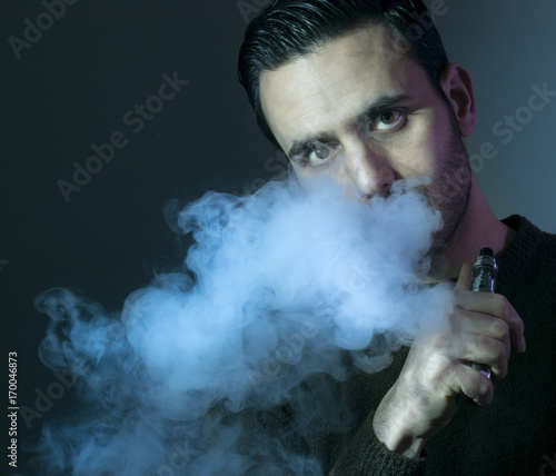 Isolated young man on a dark background holding an electronic cigarette, vaping device, mod, e-cig.
