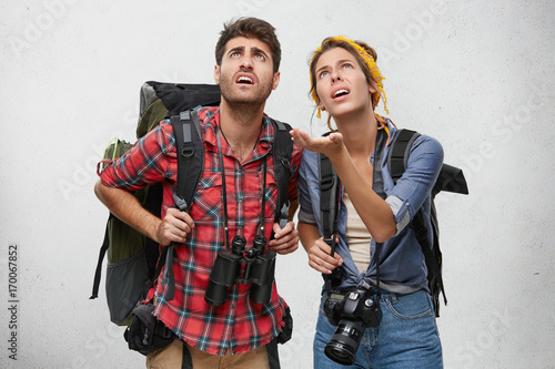 Just look at this! Astonished female with rucksack and camera showing something with hand to her husband, being shocked together, isolated over white background. Young travelers with shocked look
