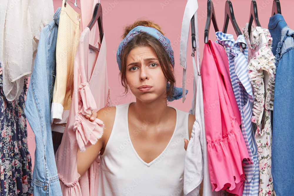 Depressed female standing near wardrobe rack full of clothes, having  difficult choice not knowing what to put on. Nothing to wear concept.  Choosing clothes for special occasion. Searching for clothing Photos