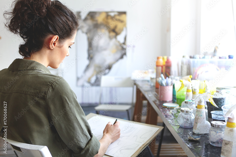 Rear view of brunette young Caucasian woman artist in khaki shirt holding pencil, sketching at workshop with paints on table near her. Art, creativity, painting, hobby, job and occupation concept Rear view of brunette young Caucasian woman artist in khaki shirt holding pencil, sketching at workshop with paints on table near her. Art, creativity, painting, hobby, job and occupation concept