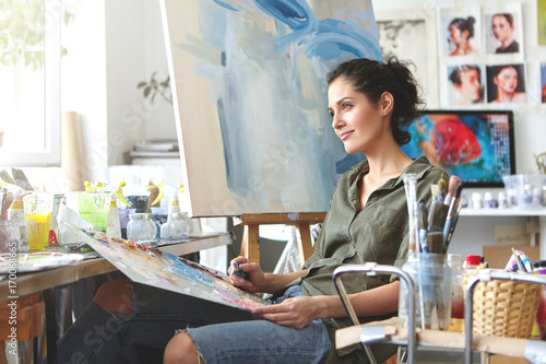 Thoughtful dreamy young European female artist bringing her creativity to life, sitting in her modern workshop interior with palette and painting knife. Hobby, job, occupation, art and craft concept photo