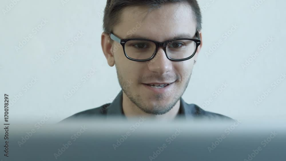 Close-up portrait of a young man wearing glasses sitting in his office in front of a monitor - having a video chat. People stock footage slider shot. Close-up portrait of a young man wearing glasses sitting in his office in front of a monitor - having a video chat. People stock footage slider shot.