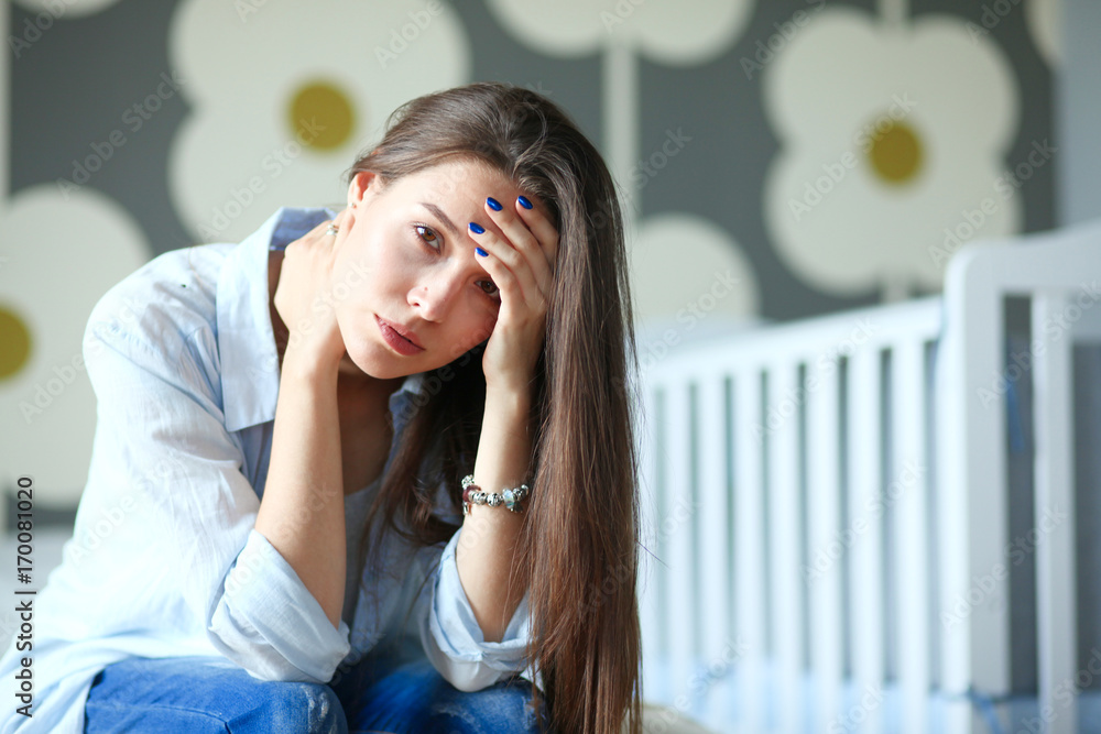 Young tired woman sitting on the bed near children's cot. Young mom. Young tired woman sitting on the bed near children's cot. Young mom.