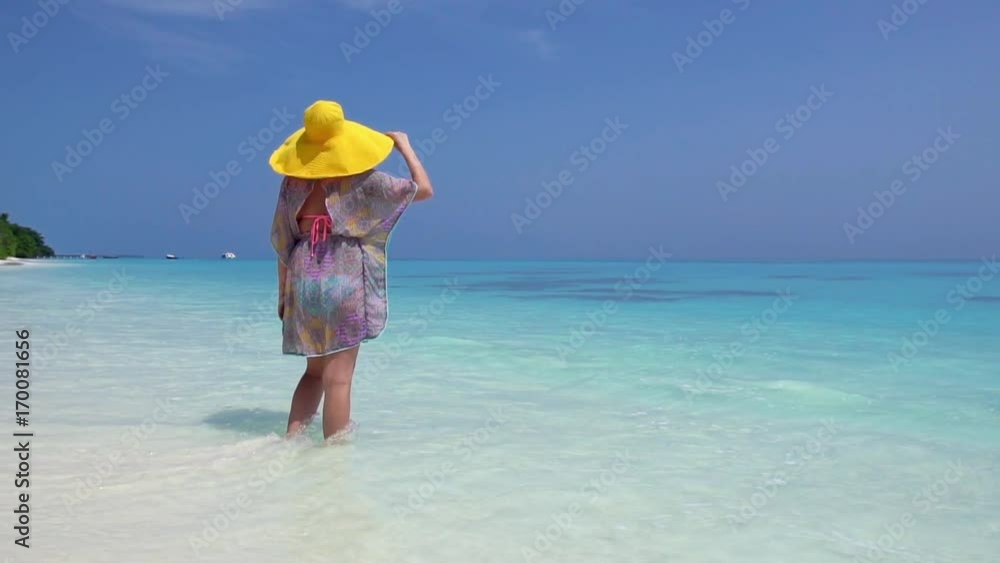 Woman in straw yellow sunhat walking along the beach with turquoise sea water Woman in straw yellow sunhat walking along the beach with turquoise sea water