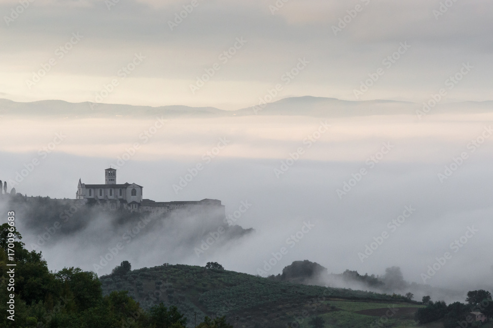 Beautiful view of St.Francis church in Assisi (Umbria, Italy), over a sea of fog at dawn, with hills and trees in the foreground