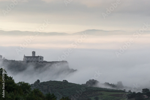 Beautiful view of St.Francis church in Assisi (Umbria, Italy), over a sea of fog at dawn, with hills and trees in the foreground