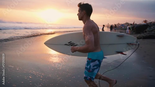Young surfer holding board, running to go in the water at amazing golden light sunset. Happy man enjoying summer evening and seaside vacation activities. Shot taken by a handheld gimbal in 60FPS