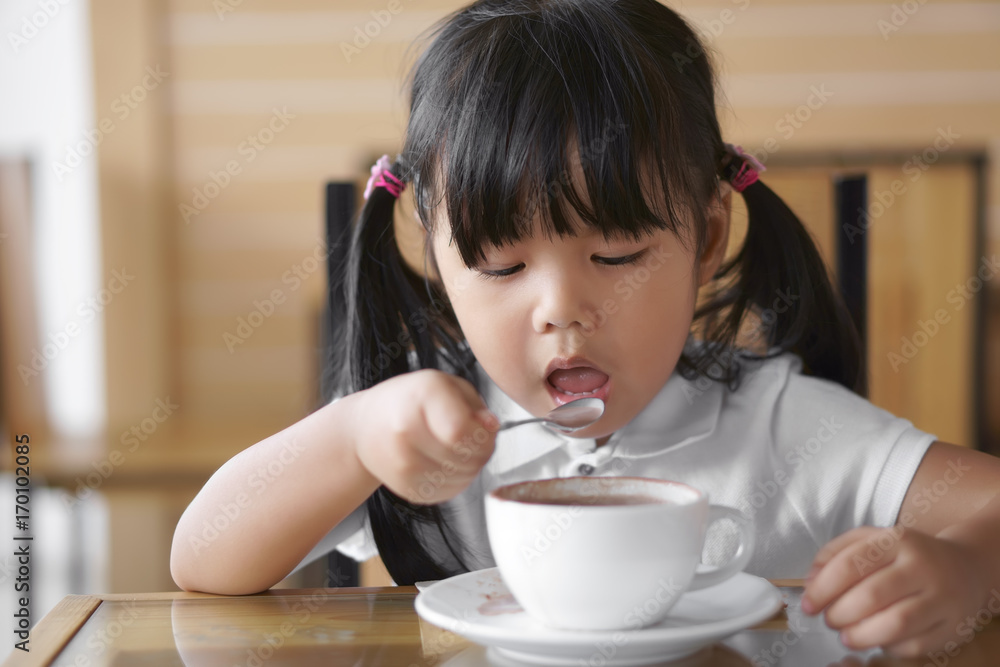 Asian children cute or kid girl holding spoon and open mouth for delicious hot cocoa or chocolate drinking in white cup and sloppy for breakfast in the morning on table in home or cafe restaurant Asian children cute or kid girl holding spoon and open mouth for delicious hot cocoa or chocolate drinking in white cup and sloppy for breakfast in the morning on table in home or cafe restaurant