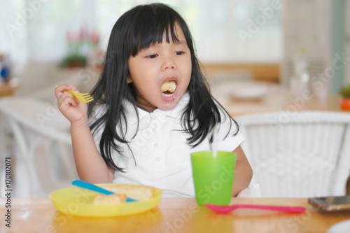 Asian children cute or kid girl enjoy eating dessert and open mouth for eat delicious bread with colorful container or utensil plastic for baby on wood table in cafe restaurant for breakfast and lunch
