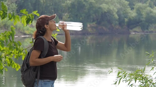 Girl traveler drinks water on the river bank. A girl with a backpack is drinking water. photo