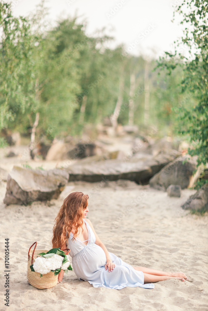 beautiful pregnant girl with long hair walking near the shore of a lake with flowers in a summer dress