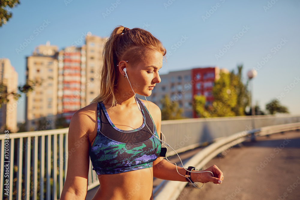 Fototapeta premium A happy girl jogger listens to music on headphones on a road in the summer city of the city.