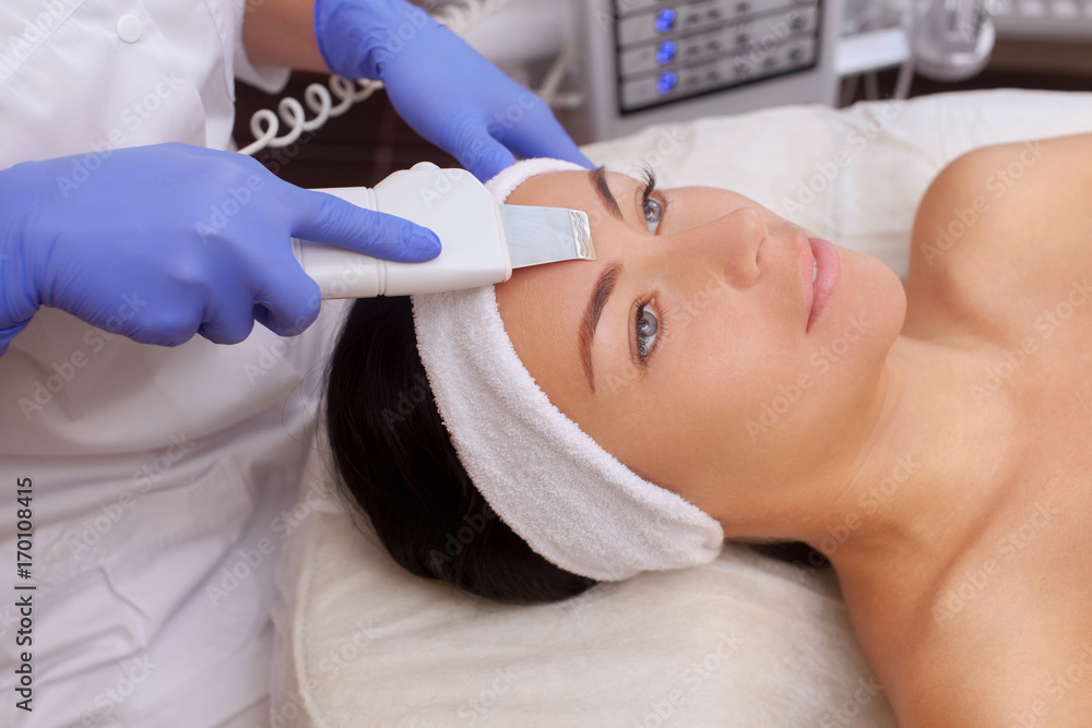 Naklejka premium The doctor-cosmetologist makes the apparatus a procedure of ultrasound cleaning of the facial skin of a beautiful, young woman in a beauty salon. Cosmetology and professional skin care.