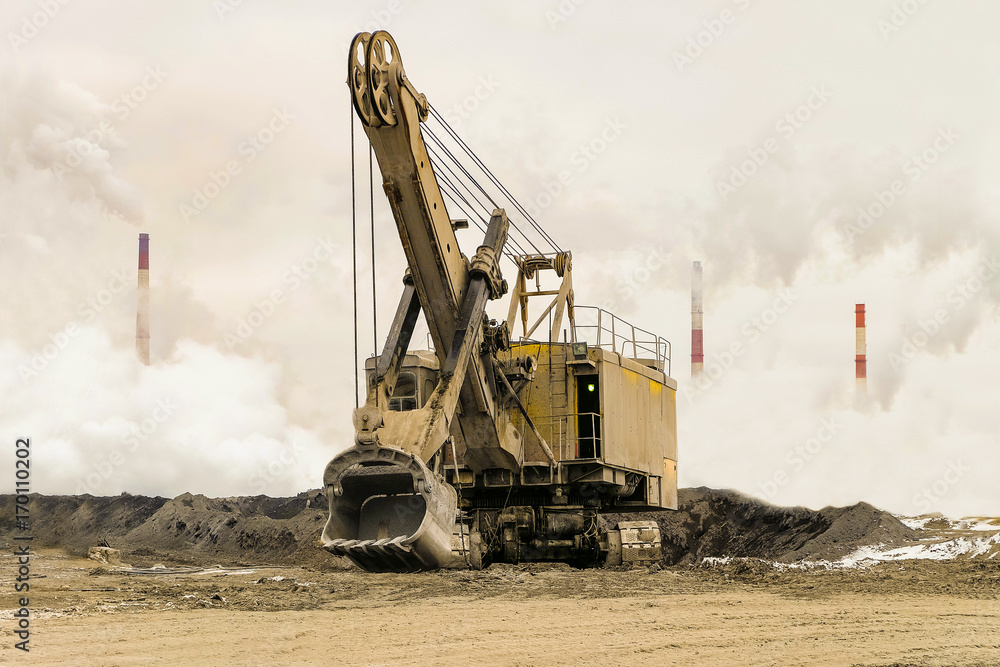 Bucket excavator on crawler track against background of dense factory smog with smoking chimneys. Heavy industry background. Excavator with mechanical drive and flexible suspension of work equipment. Bucket excavator on crawler track against background of dense factory smog with smoking chimneys. Heavy industry background. Excavator with mechanical drive and flexible suspension of work equipment.