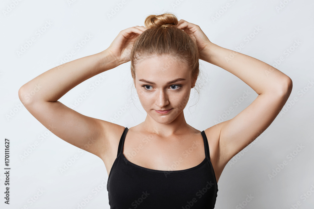 Horizontal portrait of attractive female with blonde hair knot, looking pensively down with her blue eyes while trying to make bun, thinking over where to go for walk with friends. Beauty concept Horizontal portrait of attractive female with blonde hair knot, looking pensively down with her blue eyes while trying to make bun, thinking over where to go for walk with friends. Beauty concept