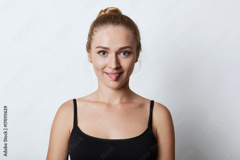 Indoor shot of funny blonde female with hair knot, showing tongue while arguing with someone, trying to persuade that she is right. Beautiful female model showing tongue into camera isolated on white Indoor shot of funny blonde female with hair knot, showing tongue while arguing with someone, trying to persuade that she is right. Beautiful female model showing tongue into camera isolated on white
