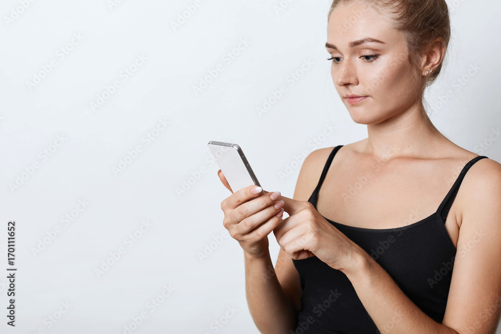 Adorable female with tied hair, wearing black casual clothes, holding modern mobile phone in hand, reading attentively electronic book. Female student preparing for final exams using smart phone Adorable female with tied hair, wearing black casual clothes, holding modern mobile phone in hand, reading attentively electronic book. Female student preparing for final exams using smart phone