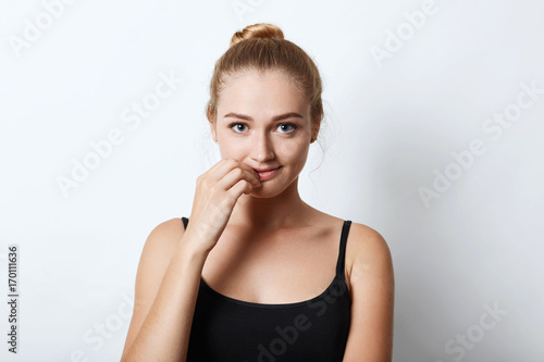 Attractive female with blue eyes, having light hair tied in hair knot, keeping hand near mouth, looking confidently into camera while wanting to say something. People, youth and beauty concept