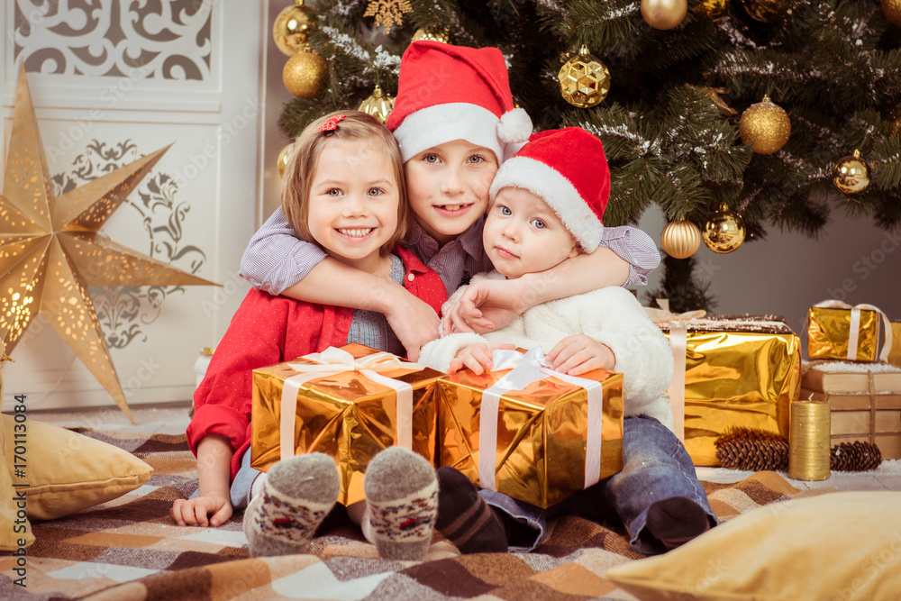 Three children in hats of Santa Claus sitting with gifts and rejoice Three children in hats of Santa Claus sitting with gifts and rejoice