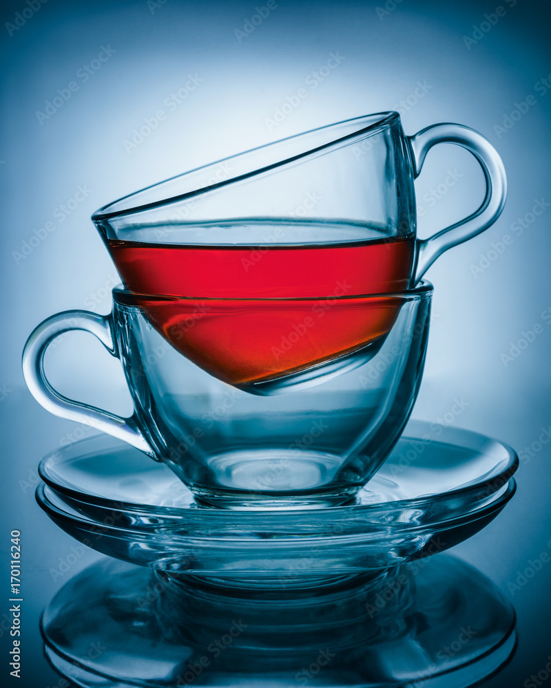 Two cups of transparent glass one inside the other on two glass plates at the top of the Cup poured tea, the whole composition is on a blue background and reflected on the surface Two cups of transparent glass one inside the other on two glass plates at the top of the Cup poured tea, the whole composition is on a blue background and reflected on the surface