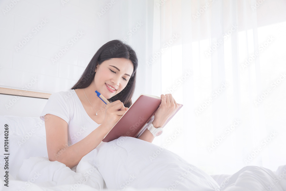 Beautiful asian woman smiling while writing diary on notebook in bedroom Beautiful asian woman smiling while writing diary on notebook in bedroom