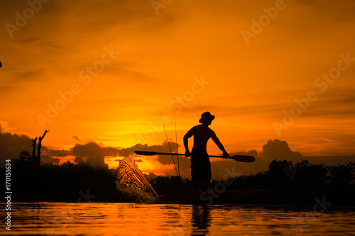 Silhouette of fisherman standing on boat,hold paddle and paddle with a splash of water on sunset background.
