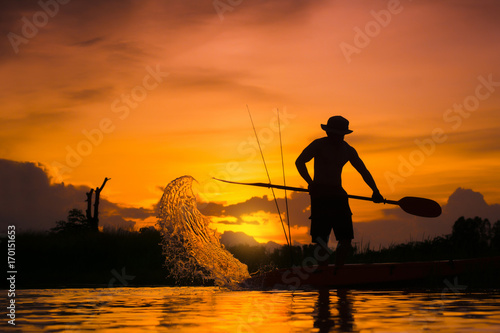 Silhouette of fisherman standing on boat,hold paddle and paddle with a splash of water on sunset background.
