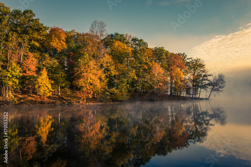 Foggy early morning sunrise over still lake with autumn trees and reflections