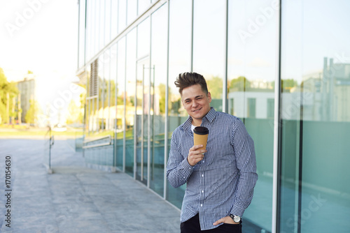 Outdoor pirture of successful trendy looking young businessman having coffee break, enjoying takeaway coffee, standing outside modern office building with glass windows, having cheerful look photo