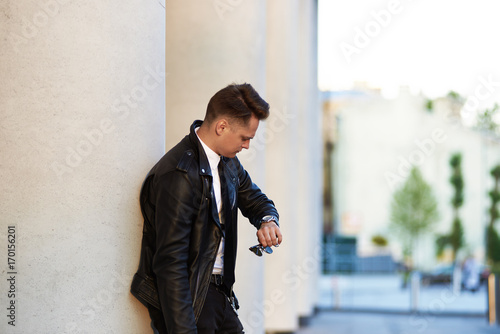 Picture of serious confident young man in stylish clothes leaning on white column outdoors and looking at his watch, checking time, getting annoyed while waiting for his girlfriend who is being late