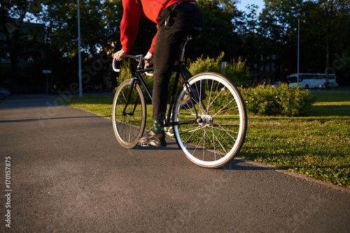 Cropped rear view of unrecognizable male in jeans and sneakers cycling along concrete path in park on fixed gear bike. Close up of white wheels of retro bicycle with stylish man riding it on sunny day © Anatoliy Karlyuk