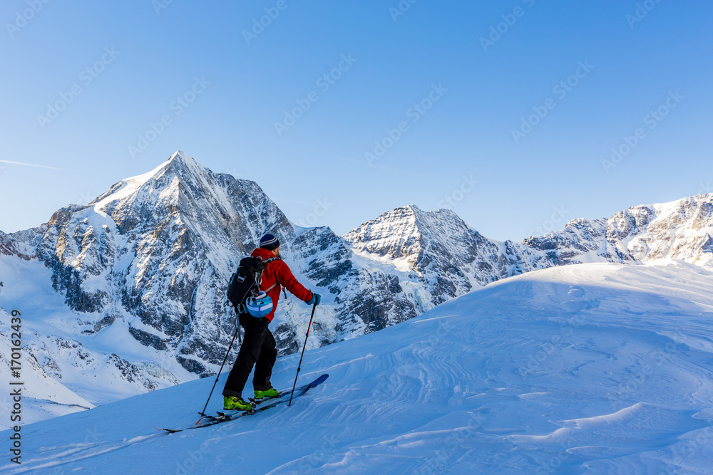 Mountaineer backcountry ski walking up along a snowy ridge with skis in the backpack. In background blue sky and shiny sun and Zebru, Ortler in South Tirol, Italy.  Adventure winter extreme sport.