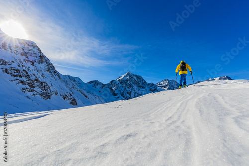 Fototapeta Mountaineer backcountry ski walking up along a snowy ridge with skis in the backpack. In background blue sky and shiny sun and Zebru, Ortler in South Tirol, Italy.  Adventure winter extreme sport.