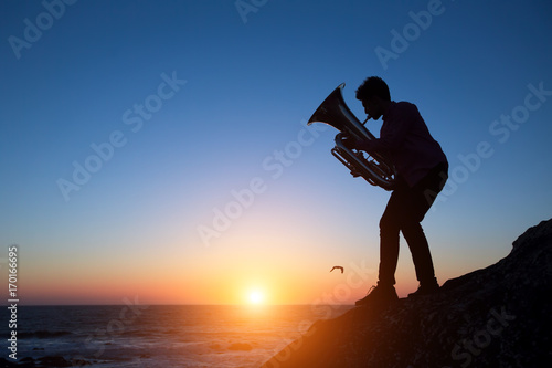 Silhouette of musician play Tuba on sea shore at amazing sunset .