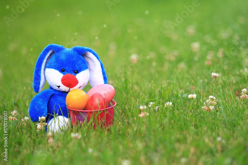 Blue bunny sitting in the grass with a basket of colorful easter eggs.